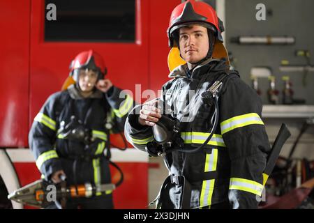 Zwei Feuerwehrleute in Schutzkleidung, Helmen und Rettungswerkzeug gegen Feuerwehrfahrzeuge Stockfoto