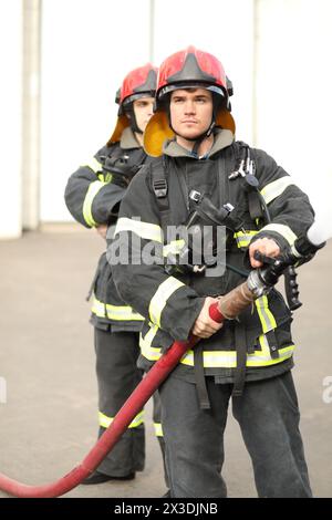 Porträt von zwei heldenhaften Feuerwehrleuten, die Düse und den Feuerschlauch halten und Hochdruckwasser spritzen, einer ist hinter dem anderen Stockfoto