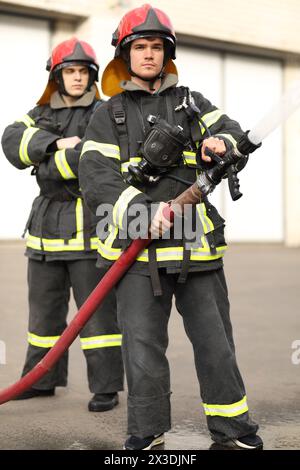 Porträt von zwei heldenhaften Feuerwehrleuten, die Düse und den Feuerwehrschlauch halten und einstellen und Hochdruckwasser spritzen Stockfoto