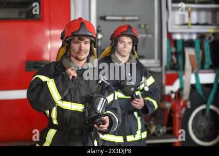 Zwei Feuerwehrmänner in Schutzanzügen und rotem Feuerwehrhelm zum Schutz des Kopfes, der gegen das Feuerwehrfahrzeug steht Stockfoto