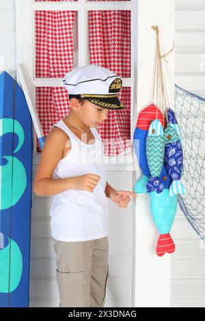 Kleiner Admiral in weißer Mütze und T-Shirt, der am Fenster neben farbigen Textilfischen steht Stockfoto