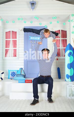 Vater in Kappe mit der Inschrift Pacific Fleet with Son in gestreiften Westen leistet akrobatische Unterstützung auf den Wappen auf der Veranda des Weißen Hauses mit Flieder Do Stockfoto