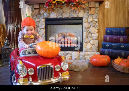 Ein kleiner Junge in einer orangefarbenen Kappe in einem roten Auto trägt einen großen Kürbis auf der Motorhaube eines Autos vor einem Kamin Stockfoto