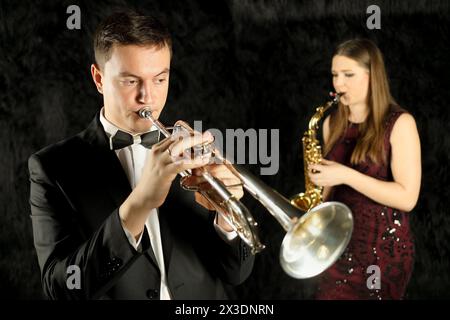 Musiker, der Trompete vor einer Frau mit Saxophon in einem schwarzen Zimmer spielt Stockfoto