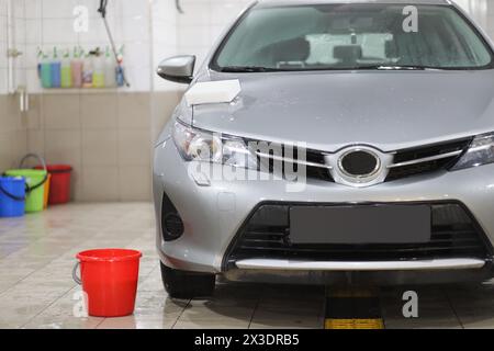 Roter Eimer mit Wasser in der Nähe eines nassen silbernen Autos und einem Schwamm auf der Motorhaube in einer Autowaschanlage Stockfoto