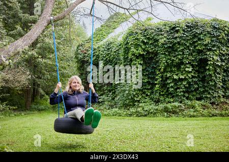 Lächelnde blonde Frau auf Seilschaukeln, die am Baum auf dem Rasen befestigt sind. Stockfoto