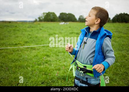 Kleiner Junge, der für Skysurfing-Stände ausgerüstet ist und fliegende Linien in der Hand auf grüner Wiese hält. Stockfoto
