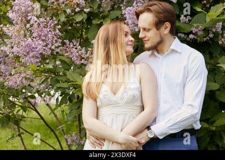 Junge lächelnde bärtige Mann und Frau stehen im Park in der Nähe des blühenden Fliederstrauchs. Stockfoto