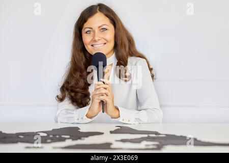 Die junge lächelnde Frau sitzt am Schreibtisch mit der Weltkarte des Puzzles und hält ein Mikrofon. Stockfoto