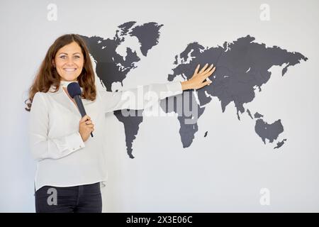 Lächelnde Frau mit Mikrofon in der Nähe der Wand mit schematischer Weltkarte. Stockfoto