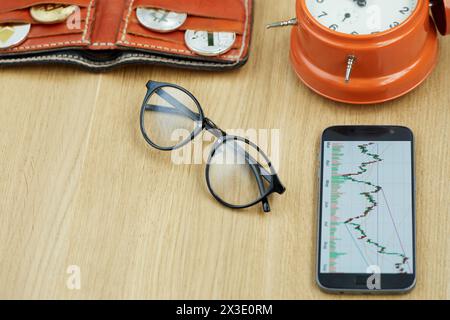 Brille, Smartphone mit Finanzkarten, Brieftasche mit Kryptomünzen, orangene Alarmuhr auf Holztisch. Stockfoto