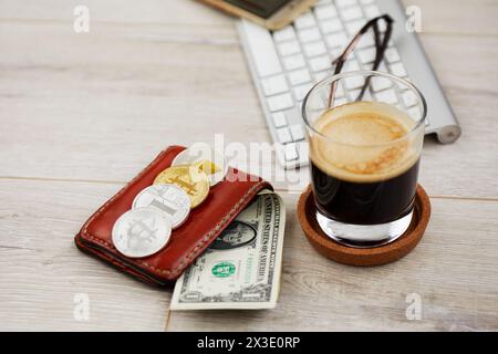 Glas Kaffee, Geldbörse mit Banknoten und Kryptomünzen, Smartphone mit Karten und Gläsern auf der Tastatur, Fokus auf Geldbörse. Stockfoto