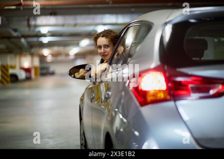 Eine junge lächelnde Frau schaut aus dem Fenster des silbernen Autos in die Tiefgarage. Stockfoto