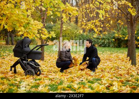 Mann und Frau sitzen mit Armvoll gefallener Blätter in der Nähe von Perambulator im Herbstpark. Stockfoto