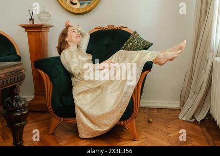 Die reife Frau in einem schönen beigefarbenen Kleid sitzt elegant auf einem grünen Sessel Stockfoto