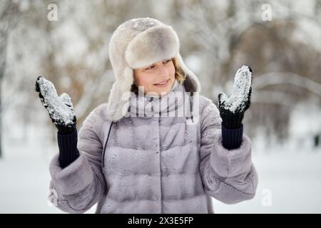 Lächelnde Frau im Pelzmantel und Hut steht und sieht im Winterpark Fäustlinge an den Händen mit festsitzendem Schnee. Stockfoto