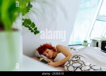 Junge rothaarige Frau liegt im Bett unter der Decke, nutzt Smartphone, Morgenferienkonzept Stockfoto