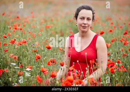 Lächelnde Frau in rotem Kleid sitzt auf rotem Mohnfeld. Stockfoto