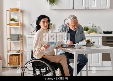 Ein Mann im Rollstuhl liest eine Zeitung, während seine Frau zu Hause an einem Tisch in ihrer Küche sitzt. Stockfoto