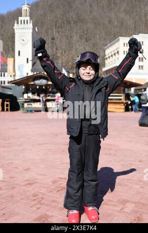 Junge in Helm und Skianzug steht mit erhobenen Armen und geschlossenen Augen auf Platz im Skigebiet mit Bergen, Krasnaja Polyana, Sotschi, Russland Stockfoto