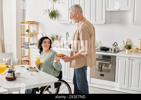 Eine Frau im Rollstuhl hält einen Teller Croissant, während sie mit seinem Partner in einer gemütlichen Küche zu Hause interagiert. Stockfoto