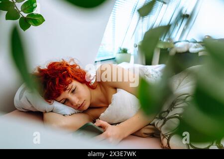 Junge rothaarige Frau liegt im Bett unter der Decke, nutzt Smartphone, Morgenferienkonzept Stockfoto