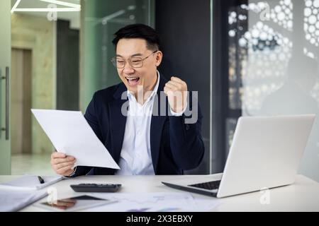 Fröhlicher asiatischer Geschäftsmann, der in einem modernen Büro mit einem Laptop Dokumente an seinem Schreibtisch liest. Stockfoto