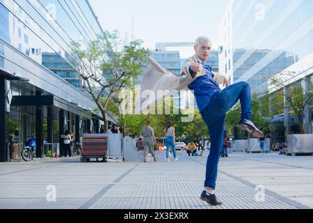 Junger schwuler Mann mit kurzen blonden Haaren, im Profil, der auf der Straße springt, sein Bein hochhebt und seine Jacke mit den Händen zurückwinkt, in die Kamera schaut, Leben Stockfoto