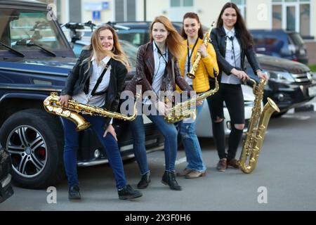 Vier Frauen posieren mit Blasinstrumenten im Freien in der Nähe von Autos, konzentrieren sich auf die linke Frau Stockfoto