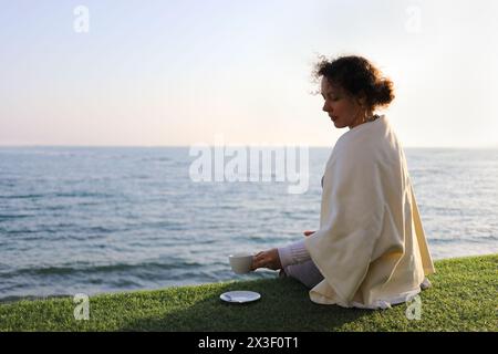 Frau in Karo sitzt auf Rasen in der Nähe des Seeufers mit einer Tasse Kaffee an sonnigem Tag Stockfoto