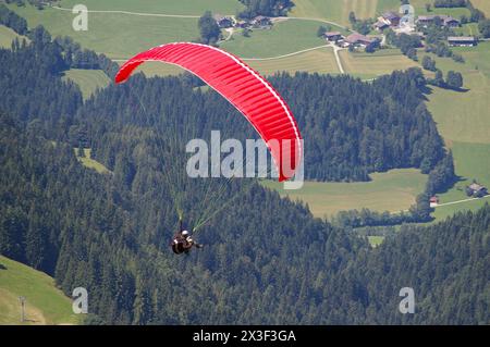 Ein roter Gleitschirmflieger, der über Wiesen- und Waldlandschaft unter sich fliegt. Stockfoto