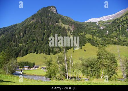 Häuser an einem Fluss, mit Bäumen bedeckten Hügel mit Wasserfall und schneebedeckten Bergen hinter einem blauen Himmel. Stockfoto