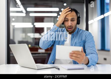 Schockierter junger hispanischer Mann, der im Büro sitzt und Kopfhörer trägt und überrascht auf den Tablet-Bildschirm blickt, der seinen Kopf mit der Hand hält. Stockfoto