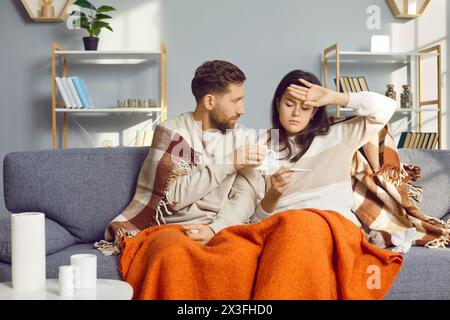 Ungesunde Mann und Frau sitzen zu Hause im Wohnzimmer, in warme Decke gewickelt und fühlen Grippesymptome. Ein junges Paar schaut sich ein Thermometer an Stockfoto