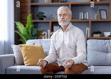 Senior entspannter grauhaariger Mann sitzt zu Hause auf dem Sofa mit geschlossenen Augen in Lotusposition, Meditation und Ruhe. Stockfoto