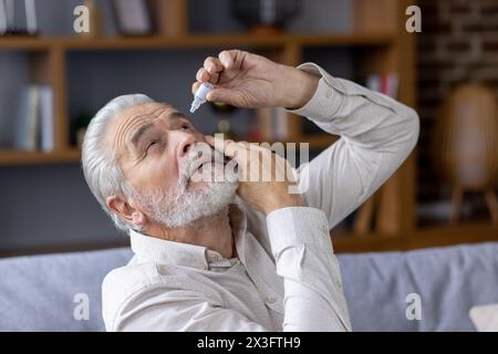 Nahaufnahme eines älteren grauen Mannes, der Augentropfen gegen Schmerzen und Erschöpfung tropft und zu Hause auf der Couch sitzt. Stockfoto