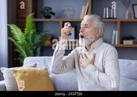 Ein älterer grauhaariger Mann leidet an Asthma. Er sitzt zu Hause auf der Couch und benutzt einen Inhalator. Stockfoto