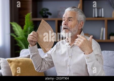 Nahaufnahme eines grauhaarigen älteren Mannes, der zu Hause auf dem Sofa sitzt und an Schlaflosigkeit leidet, einen Ventilator in der Hand hält und im Gesicht winkt. Stockfoto