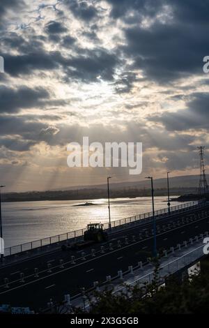 Dramatischer Himmel mit Sonnenstrahlen, die Wolken über einer ruhigen Küstenstraße in der Abenddämmerung durchdringen Stockfoto