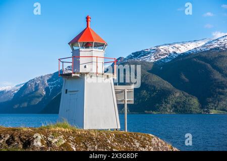Leuchtturm auf felsigem Hügel entlang der Küste der malerischen norwegischen Stadt, entlang des norwegischen Sognefjords mit blauem Himmel, kleinen Wolken und grünen schneebedeckten Bergen Stockfoto