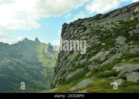 Ein hoher Berg mit einem steilen, mehrschichtigen Hang, der mit Gras bedeckt ist und an einem sonnigen Sommertag Felsformationen überblickt. Ergaki Naturpark, Krasnoyars Stockfoto