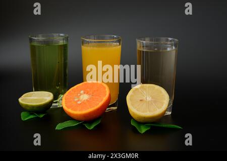 Drei große Gläser mit verschiedenen Zitrussäften auf schwarzem Hintergrund, neben den Hälften reifer Zitrone, Orange und Limette mit Blättern. Nahaufnahme. Stockfoto