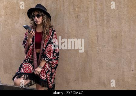 Junge Latina-Frau mit lockigen Haaren, Hut, Sonnenbrille, die auf ihrem Handy spricht Stockfoto