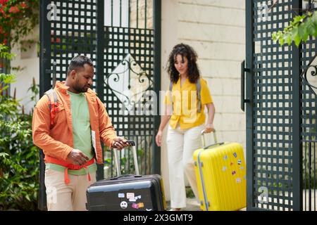 Junge interkulturelle Paare in Casualwear verlassen ihr Haus und schieben Koffer mit Gepäck, bevor sie ein Taxi nehmen und zum Flughafen fahren Stockfoto
