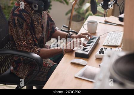 Zugeschnittene Aufnahme eines jungen kreativen Mannes, der im Heimstudio am Schreibtisch sitzt und während der Musikaufnahme die Tasten des elektronischen Synthesizers drückt Stockfoto