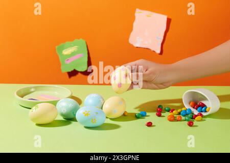 Einige Ostereier in pastellblauer und gelber Farbe sind auf grüner Oberfläche mit Süßigkeiten und einer Schale mit Farbtönen dekoriert. Papierstücke mit Zettel Stockfoto