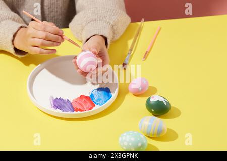 Eine Frau, die einen Pullover trägt, malt ein Osterei mit einem Pinsel, auf der Schale waren drei Farben. Ein paar Ostereier mit verschiedenen Patte Stockfoto