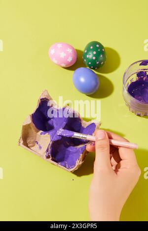 Eine weibliche Hand malt einen Eierbehälter mit violetter Farbe, andere Ostereier sind auf der Oberfläche angeordnet. Der Ostersonntag ist einer der festlichsten Stockfoto