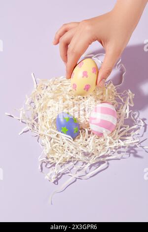Eine Menge Kraftpapier auf lila Hintergrund mit bunten Ostereiern als Nest verziert. Eine Hand nimmt ein Ei auf, das mit rosa Häschen bemalt ist. Konz Stockfoto