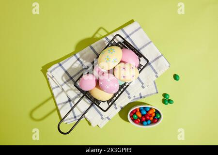 Konzept des Ostertags mit der Komposition eines Korbs mit vielen bunten Ostereiern auf gefaltetem Handtuch und einer kleinen Schüssel Süßigkeiten. Stockfoto